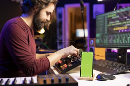 Music producer uses controls and knobs on mixing console in home studio, having a smartphone with greenscreen display. Artist learning to edit tunes and add sound effects with daw software.