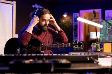 Sound composer operating with electronic setup in home studio, recording and playing acoustic guitar tunes. Musician working with signal processing techniques and audio plug ins at desk.