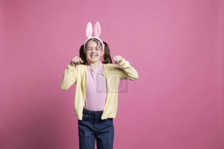 Photo for Delightful child imitates a rabbit for the Easter celebration, hopping in the studio while she is wearing bunny ears. Cheerful young kid jumping against pink backdrop, acting goofy and amusing. - Royalty Free Image