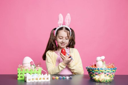 Téléchargez les photos : Jolie petite fille colorant oeufs avec aquarelle et pinceau pour les préparations de Pâques, décoration ornements festifs sur fond rose. Petit bambin s'amuser avec des décorations. - en image libre de droit