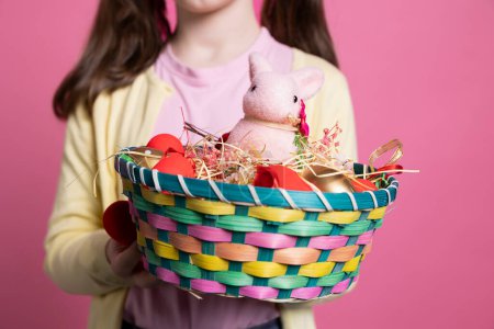 Photo for Small child presenting easter decorations and toys in a basket, holding colorful spring festive arrangements in front of camera. Young positive girl smiling in studio with adorable decor. Close up. - Royalty Free Image