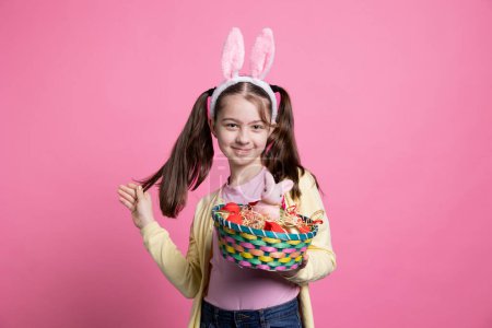 Téléchargez les photos : Petit enfant aux oreilles de lapin tient jouets et décorations de Pâques, évoquant l'enchantement de la saison. Jolie fille avec un sourire adorable montrant un lapin et des œufs dans un panier sur la caméra. - en image libre de droit