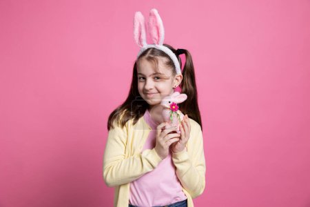 Photo for Positive cheerful girl with bunny ears posing over pink background, feeling enthusiastic about easter festive celebration. Cute small child holding a pink rabbit toy in front of camera, fluffy items. - Royalty Free Image