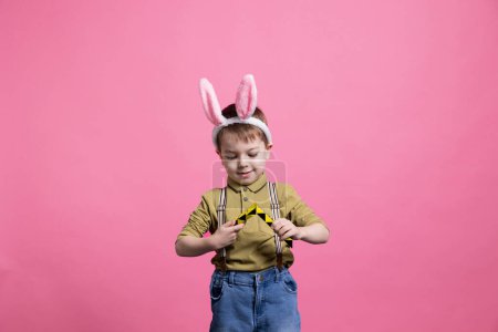 Téléchargez les photos : Joyeux petit enfant jouant avec un jouet coloré devant la caméra, profitant d'une activité de loisirs sur fond rose. Jeune garçon joyeux portant des oreilles de lapin et posant pour une séance photo de Pâques. - en image libre de droit