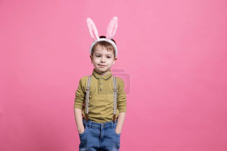 Téléchargez les photos : Jeune enfant joyeux portant de jolies oreilles de lapin devant la caméra, se sentant excité par la fête de Pâques et debout sur fond rose. Petit garçon joyeux avec une adorable tenue. - en image libre de droit