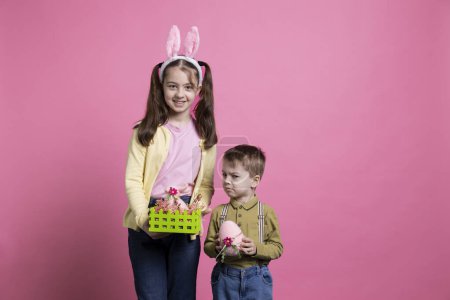 Téléchargez les photos : Enfants ludiques montrant des œufs peints et décorant pour la fête de Pâques, debout ensemble sur fond rose. Frère et sœur se sentent joyeux et enthousiastes à propos de l'événement d'avril. - en image libre de droit