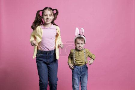Téléchargez les photos : Frère avec des oreilles de lapin et sœur sautant dans le studio, les petits enfants étant heureux de la célébration de Pâques. Adorables frères et sœurs rebondissant comme des lapins sur fond rose. - en image libre de droit