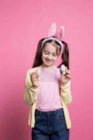 Photo for Small child posing with confidence in front of camera, showing her painted colorful eggs for easter celebration. Young sweet kid with bunny ears smiling in studio, presenting handmade decorations. - Royalty Free Image