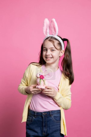 Photo for Cheerful small kid posing with a pink rabbit toy on camera, wearing fluffy bunny ears and holding easter ornaments over background. Young toddler with pigtails being excited about festive event. - Royalty Free Image