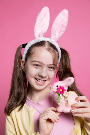 Photo for Joyful pretty toddler with bunny ears and pigtails holding a pink rabbit toy, posing with confidence over pink background. Young girl feeling optimistic about easter celebration, colorful decor. - Royalty Free Image