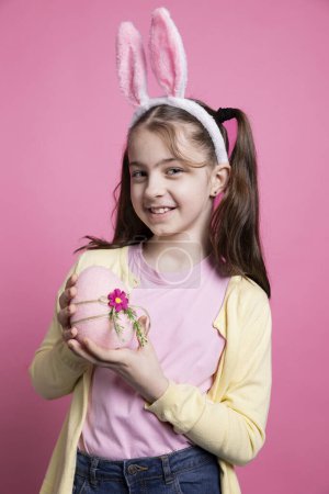 Photo for Happy positive girl with bunny ears holds a pink easter egg in studio, feeling proud about her festive decorations handmade. Small child being confident on camera, spring holiday event. - Royalty Free Image