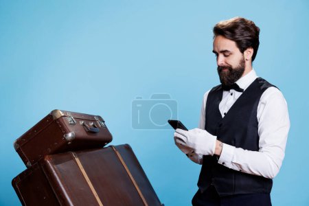 Young bellboy surfing web on smartphone, acting like a professional luxury hotel employee on camera. Doorman using internet sites, reviewing online bookings or services reservations.