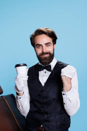 Confiado portero sintiéndose feliz en el estudio, sosteniendo la taza de café sobre fondo azul. Sonriente empleado de botones con un atuendo elegante y beber cerveza fría de refresco de té.