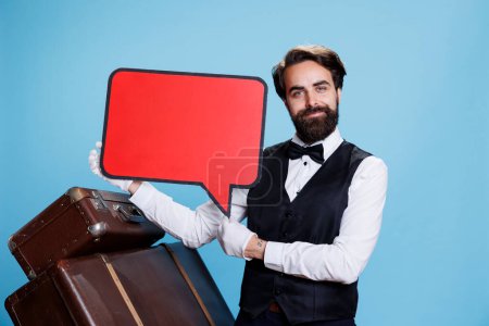 Hotel worker showing ad with speech bubble, holding empty isolated red billboard icon. Elegant doorkeeper presents blank copyspace cardboard sign on camera against blue background.