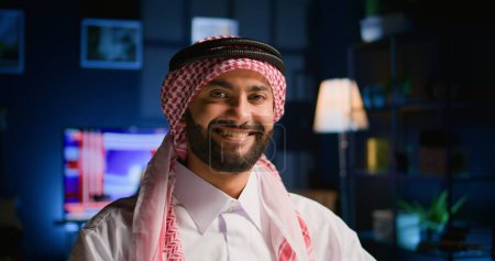Photo for Portrait of happy arabic teleworker sending job emails in stylish apartment. Cheerful muslim employee remotely working, typing data on laptop with opened tv as background noise, close up shot - Royalty Free Image