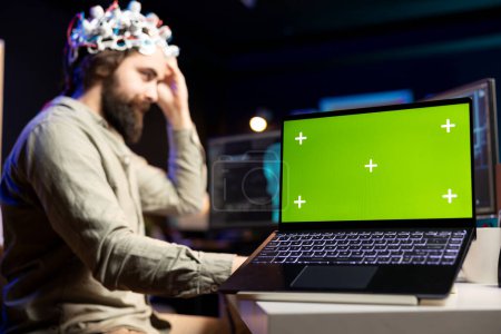 Téléchargez les photos : Concentrez-vous sur l'ordinateur portable à écran vert utilisé par le développeur portant un casque EEG traduisant les pensées en commandes PC. Professionnel de l'informatique de contrôle des fonctions informatiques en utilisant l'esprit, aidé par ordinateur portable maquette - en image libre de droit