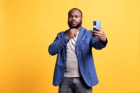 Photo for Narcissistic man using cellphone to take selfies, stroking his chin. Vain social media user taking photos using phone selfie camera, isolated over yellow studio background - Royalty Free Image