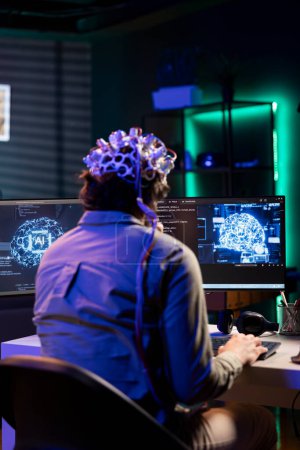 Téléchargez les photos : Technicien logiciel avec casque EEG sur la programmation du transfert de cerveau dans le monde virtuel de l'ordinateur, fusionnant avec l'intelligence artificielle. Expert informatique utilisant les neurosciences pour gagner l'âme numérique - en image libre de droit