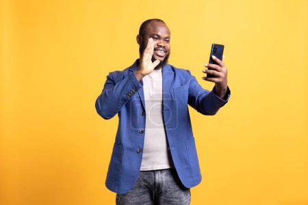 Joyful african american man greeting friends during teleconference meeting using smartphone, studio background. Happy person waving hand, saluting mate during internet video call