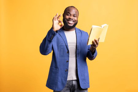 Portrait of smiling man recommending interesting book after being entertained by well written story. Joyous person showing ok hand sign, enjoying literature novel, studio background