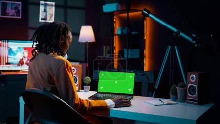 Téléchargez les photos : Une jeune femme afro-américaine regarde un ordinateur portable avec un écran vert, assise à son bureau et se préparant à suivre des cours universitaires en ligne. Fille en utilisant PC avec mise en page de maquette isolée. Caméra B. - en image libre de droit