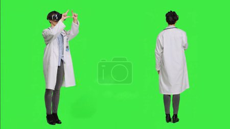 Woman medic works with 3d virtual reality headset in studio, standing against greenscreen backdrop. General practitioner uses vr glasses with interactive vision tool, futuristic exam. Camera A.