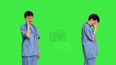 Téléchargez les photos : Infirmière malade souffrant d'un mal de tête sur fond d'écran vert, portant des blouses et se sentant surmenée à l'hôpital. Assistant médical ayant une migraine douloureuse, le stress et l'épuisement professionnel. Caméra B. - en image libre de droit