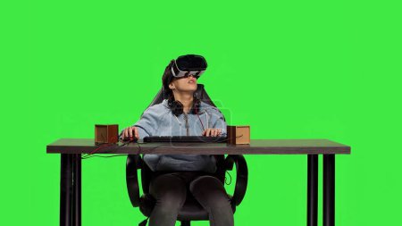 Pleased woman celebrating her video games online success at computer, enjoys playing tournament with virtual reality interactive headset. Gamer feels happy against greenscreen backdrop. Camera B.
