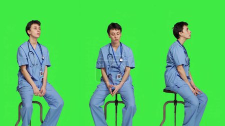 Photo for Medical assistant waiting for someone while she sits on chair, feeling impatient and waits for patients to attend checkup appointments. Nurse in scrubs against greenscreen backdrop. Camera A. - Royalty Free Image