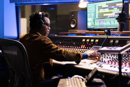 Foto de El productor de sonido afroamericano utiliza compresor y mezclador en un estudio de grabación profesional, ajustando los niveles de volumen y la configuración de audio en una pista. Concepto de producción musical en sala de control. - Imagen libre de derechos