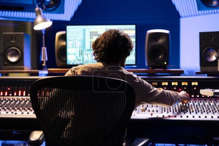 Skilled sound designer working on digital audio software app, editing tracks with mixing console and control panel board. Male engineer producing music with faders and buttons in studio.