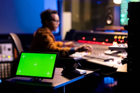Photo for African american sound engineer editing music next to greenscreen display on gadget, operating on control room mixing console. Young producer pressing knobs and sliders in studio. - Royalty Free Image