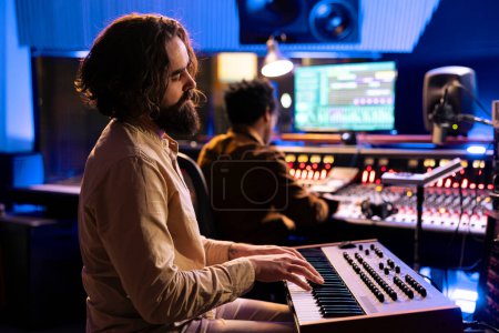 Photo for Young artist songwriter creating a new song with piano keys in studio, collaborating with audio engineer in control room. Musician singer composing music with midi controller electronic keyboard. - Royalty Free Image
