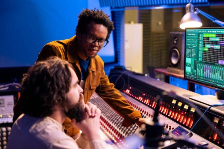 African american artist works with producer in professional studio, talking about adding new sound effects on tracks recorded. Young musician and technician editing songs with control panel desk.