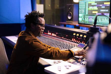 Afroamerikanischer Musikproduzent komponiert Tracks im Regieraum, schneidet Songs und passt Lautstärkeeinstellungen mit Armaturenbrettknöpfen an. Tontechniker drückt Fader und Tasten.