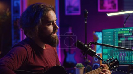 Songwriter producing tunes on acoustic guitar in his personal studio, using a microphone to sing the chords. Audio engineer works with soundboard and equalizer to compose new music. Camera A.