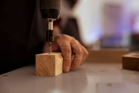 Woodworker in assembly shop using power drill to create holes for dowels in wooden board. Carpenter sinking screws into wooden surfaces with electric tool, doing precise drilling for seamless joinery