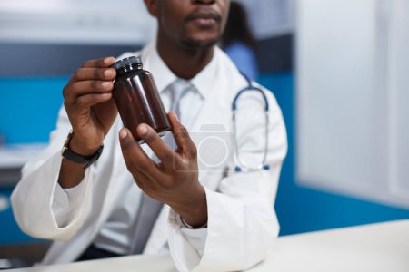 Photo for Close-up shot of a black man wearing a lab coat and holding a prescription medicine in the clinic office. Detailed image of African American doctor grasping a bottle of pills for recovery treatment. - Royalty Free Image