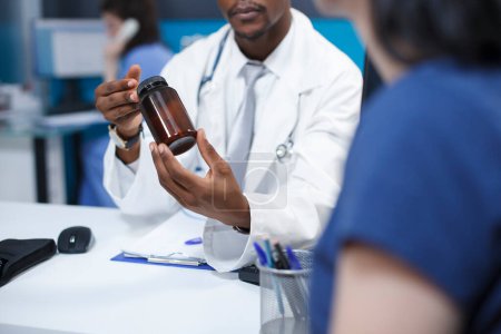 Photo for Close-up shot of an African American doctor describing the diagnosis and course of treatment of a caucasian patient while holding a bottle of medication. A woman receiving medical advice. - Royalty Free Image