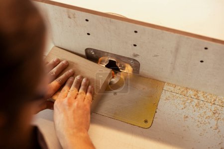 Carpenter in studio puts wood block through spindle moulder, creating smooth edges on wooden pieces. Cabinetmaker in carpentry shop using heavy machinery to craft strong seamless joints for furniture