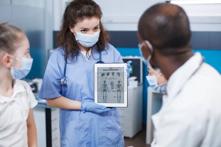Photo for During a visit, bone doctor and nurse discuss the best medical alternatives for the patient in the clinic office. A doctor in antibacterial protective gear displays orthopedic information on a tablet. - Royalty Free Image