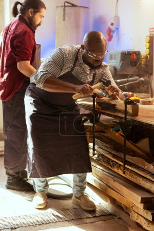 Téléchargez les photos : Fabricant afro-américain sculptant des conceptions complexes dans le bois à l'aide de ciseau et de marteau, portant des lunettes de sécurité. Artisan en atelier de menuiserie façonnant des pièces en bois à l'aide d'équipements de protection - en image libre de droit