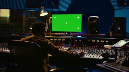 Afroamerikanischer Musikproduzent, der mit Greenscreen am PC arbeitet und das Mischpult benutzt, um den Mix und das Mastering in der Regie durchzuführen. Tontechniker nehmen Spuren mit Knöpfen und Schaltern auf. Kamera B.