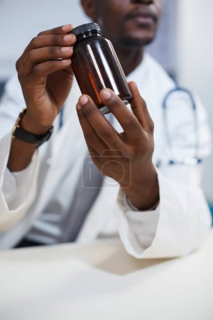 El primer plano muestra a un hombre negro usando una bata de laboratorio y agarrando una pastilla recetada. Enfoque selectivo de un médico afroamericano que sostiene una botella de medicamento para el tratamiento del paciente.