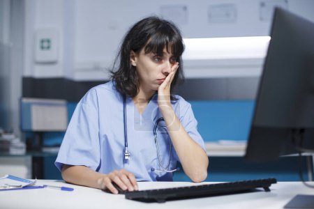 Foto de En la oficina, el asistente médico utiliza una computadora y un ratón para el sistema de salud. Mientras trabaja hasta tarde en la noche, la enfermera está mirando a la pantalla del monitor en busca de apoyo y asistencia. - Imagen libre de derechos