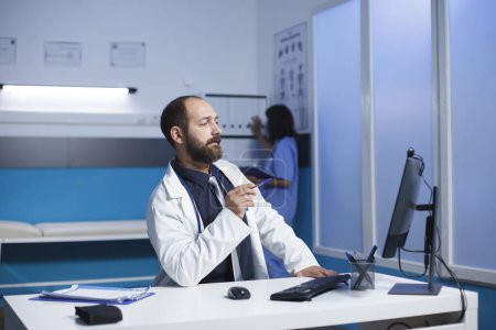 Téléchargez les photos : Docteur dans un hôpital communique par un appel vidéo à l'aide de son ordinateur de bureau. Un homme caucasien assis dans le bureau de la clinique ayant une réunion virtuelle. Collaboration et technologie dans le domaine de la santé. - en image libre de droit