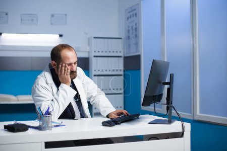 Bild, das einen müden kaukasischen Arzt zeigt, der nach einem langen Tag im Krankenhaus an einem Schreibtisch mit einem Desktop-Computer sitzt. Erschöpfter Arzt berührt sein Gesicht im Praxiszimmer einer Klinik.