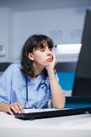 Téléchargez les photos : Femme praticienne caucasienne dans une clinique moderne, fatiguée mais concentrée. Avant un rendez-vous, une infirmière formée par un pharmacien analyse un bilan médical à l'aide d'un ordinateur dans le bureau de l'hôpital. - en image libre de droit