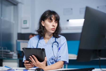 Téléchargez les photos : Infirmière caucasienne en uniforme médical analysant les prescriptions pour les patients sur tablette et ordinateur, travaillant dans un bureau d'hôpital. Assistance médicale comparant les données de santé sur les appareils numériques. - en image libre de droit