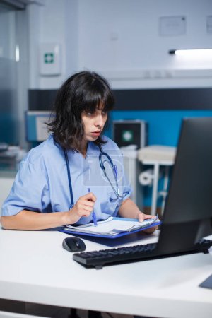Photo for Focused female nurse is examining and rereading her notes at the clinic office room. When getting ready for patient medical consultations, caucasian practitioner uses desktop monitor and clipboard. - Royalty Free Image